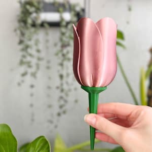 May include: A pink tulip-shaped plant waterer with a green stem. The waterer is held in front of a blurred background of greenery. The tulip head is a metallic rose colour, and the stem is a vibrant green.