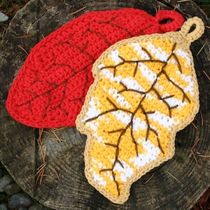 May include: Two leaf-shaped crocheted pot holders. One is solid red with brown vein detailing. The other is yellow and white with brown vein detailing, and a beige border. Both have a loop for hanging.