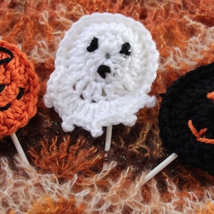 May include: Three crocheted Halloween-themed lollipops. A pumpkin, a ghost, and a black cat.