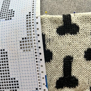 May include: Close-up of a knitted fabric with a repeating black pattern on a cream background. A knitting pattern chart is pinned to the fabric. The chart has rows and columns with punched holes. Yellow and blue pins secure the fabric.