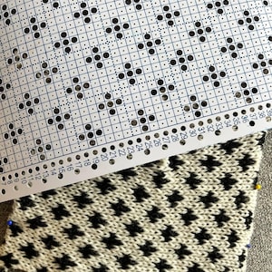 May include: Close-up of a knitting pattern and a partially completed knitted swatch. The pattern is a white card with black dots and numbers. The knitted swatch features a black and white star pattern, secured with pins.