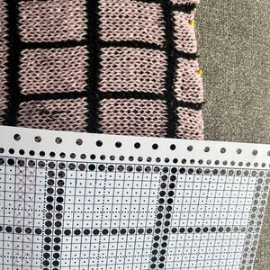 Könnte beinhalten: Nahaufnahme eines Strickstoffs mit einem schwarzen Gittermuster auf rosa Hintergrund. Der Stoff ist teilweise sichtbar, mit gelben Nadeln am Rand. Eine weiße Lochkarte mit einem Raster aus Löchern und Punkten befindet sich im Vordergrund.