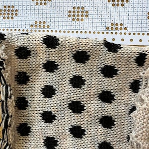 Op de afbeelding: Close-up van een gebreide stof met een crèmekleurige basis en grote zwarte stippen. De stof ligt naast een papieren patroon met een rasterontwerp. De rand van de stof heeft een franje van zwart en crèmekleurig garen.