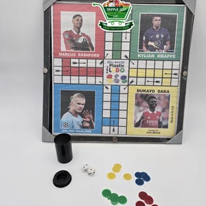 May include: A plastic Ludo board game with a black frame. The board features images of famous soccer players, including Marcus Rashford, Kylian Mbapp&eacute;, Bukayo Saka, and Erling Haaland. The game includes four colored playing pieces, two dice, and a cup.