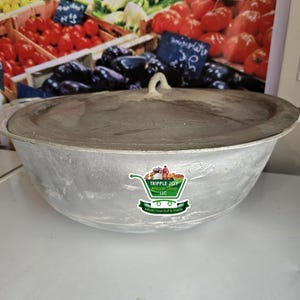May include: A large, round, silver cooking pot with a lid and a handle. The pot is sitting on a white surface. The pot has a sticker on the side that says "Tripple Joy African Store LLC".