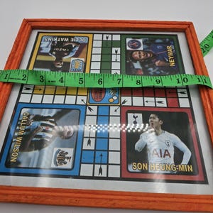 May include: A framed wooden game board with a colorful design featuring soccer players. The board has a grid pattern with arrows pointing in different directions. The players featured include Callum Wilson, Son Heung-min, Ollie Watkins, and Neymar.