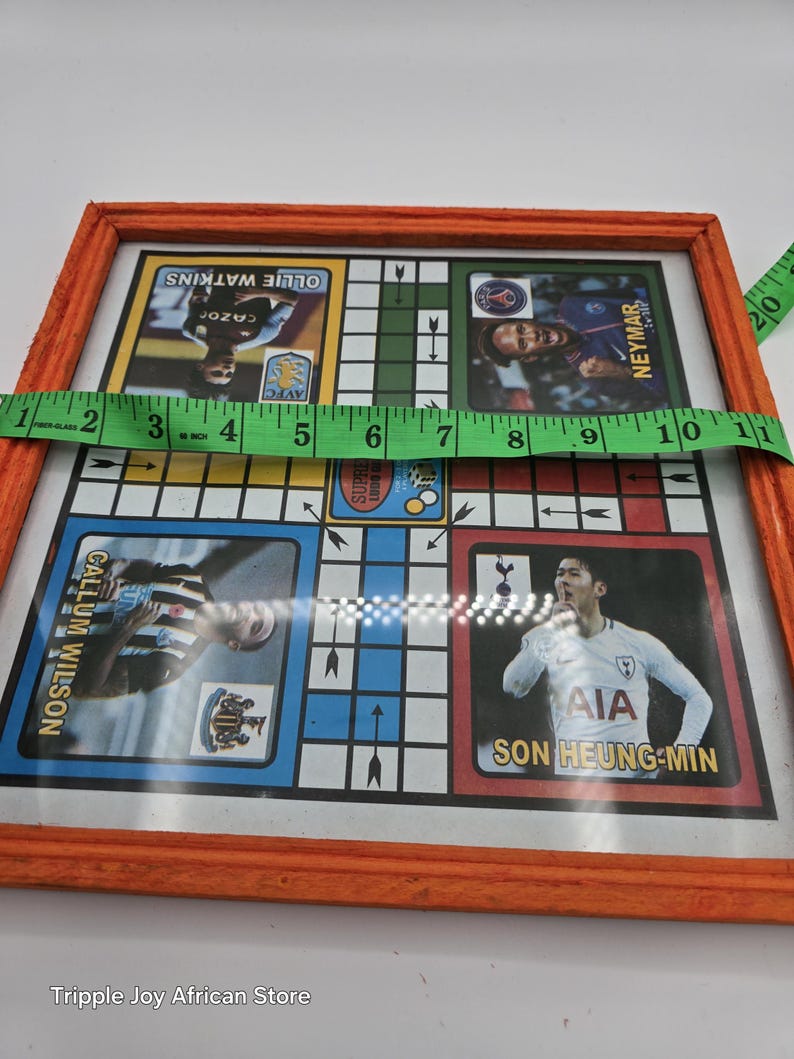 May include: A framed wooden game board with a colorful design featuring soccer players. The board has a grid pattern with arrows pointing in different directions. The players featured include Callum Wilson, Son Heung-min, Ollie Watkins, and Neymar.
