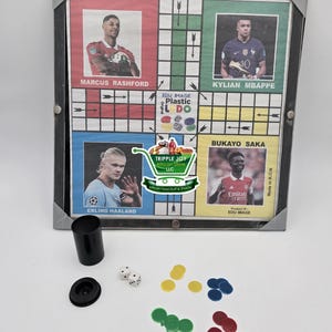 May include: A framed plastic Ludo board game with images of famous soccer players, including Marcus Rashford, Kylian Mbappe, Bukayo Saka, and Erling Haaland. The game includes dice, a cup, and colored playing pieces.