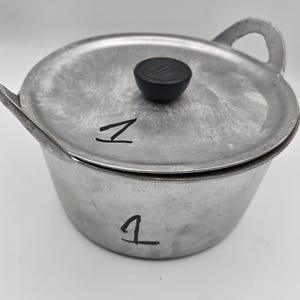 May include: A silver-colored aluminum cooking pot with a matching lid and two side handles. The lid features a black circular knob. The number 1 is handwritten in black ink on both the lid and the side of the pot. The text Tripple Joy African Store is visible.
