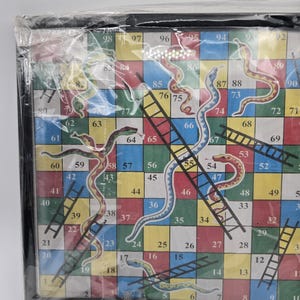 May include: A Snakes and Ladders board game, featuring a colorful grid with numbers, snakes, and ladders. The game board is in a black frame and wrapped in plastic. The game is from Tripple Joy African Store.