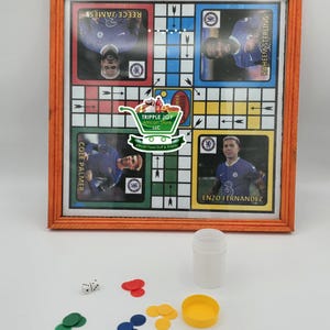 May include: A wooden framed game board with a colorful checkerboard pattern. The board features images of soccer players and the text "Cole Palmer", "Reece James", "Raheem Sterling", and "Enzo Fernandez". The game board is accompanied by a white plastic container, two white dice, and a set of colorful game pieces.
