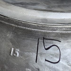 May include: Close-up of a metallic gray wheel rim. The number "15" is embossed and written on the surface. The rim appears to be part of a vehicle's wheel assembly.