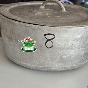 May include: A large, silver, cast iron pot with a lid and a handle. The pot has a sticker on the side that says "Tripple Joy African Store LLC" and a handwritten number "8" on the side.