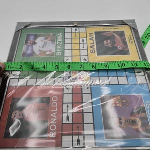 May include: A board game featuring images of famous soccer players, including Ronaldo, Benzema, Salah, and Messi. The board is made of cardboard and has a black frame. The game is made in the UK and is suitable for ages 6 and up.