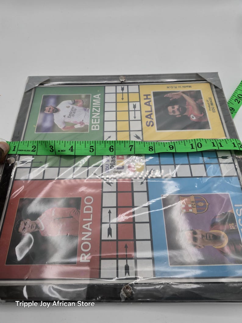 May include: A board game featuring images of famous soccer players, including Ronaldo, Benzema, Salah, and Messi. The board is made of cardboard and has a black frame. The game is made in the UK and is suitable for ages 6 and up.