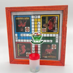 May include: A wooden framed game board with a grid of squares and images of soccer players. The board is red and has the names "Bruno Fernandes", "Marcus Rashford", "Edinson Cavani", and "Mason Greenwood" printed on it. A clear plastic container with a red lid and dice inside is in the foreground.