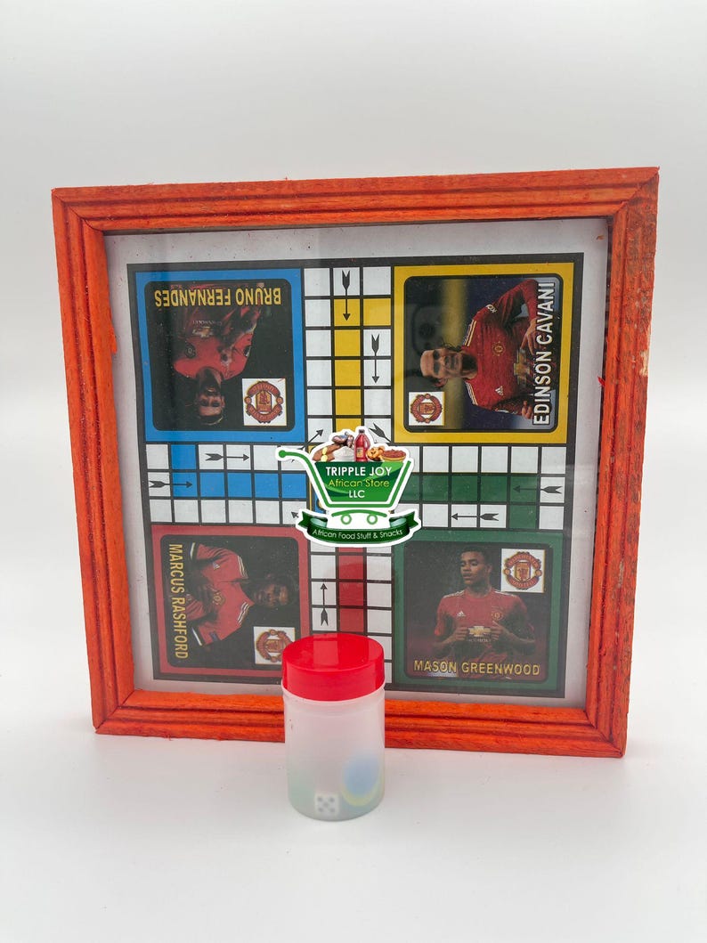 May include: A wooden framed game board with a grid of squares and images of soccer players. The board is red and has the names "Bruno Fernandes", "Marcus Rashford", "Edinson Cavani", and "Mason Greenwood" printed on it. A clear plastic container with a red lid and dice inside is in the foreground.