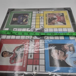 May include: A game board with a grid of squares and arrows. The board features photos of soccer players, including Kylian Mbappe, Bukayo Saka, Marcus Rashford, and Erling Haaland. The board is made of cardboard and is covered in plastic.