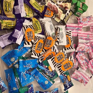 May include: A variety of individually wrapped candies in a clear plastic container. The candies are in a variety of colors and flavors, including chocolate, mint, and fruit. Some of the candies have zebra-print wrappers, while others have wrappers with the words "Tom Tom" and "Milo".