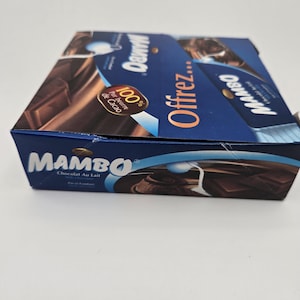 May include: A blue and white box of Mambo chocolate bars. The box features the brand name "Mambo" in large white letters and the text "Chocolat Au Lait" in smaller white letters. The box also features a picture of a chocolate bar and the text "100% Pur beurre de Cacao".