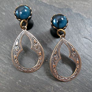 May include: A pair of ornate earrings featuring teardrop-shaped pendants with intricate patterns. Each earring has a dark teal-colored, round gem at the top, connected to the pendant by a small brass ring. The pendants are a copper color.