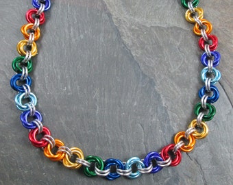 May include: A rainbow colored chainmail necklace with a silver clasp. The necklace is made of small metal rings that are linked together in a pattern. The rings are colored in a rainbow pattern, with each ring being a different color.