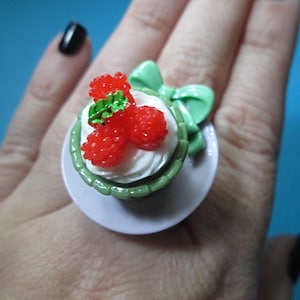 May include: A novelty ring featuring a miniature cupcake design. The ring has a white frosting base, topped with red raspberry-like beads and green leaves. A green bow is attached to the side. The ring is set on a white base.