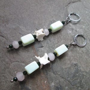 May include: A pair of earrings with a unique design featuring white bone beads, light green and pink glass beads, and black beads. The earrings are made with silver-toned metal hooks.