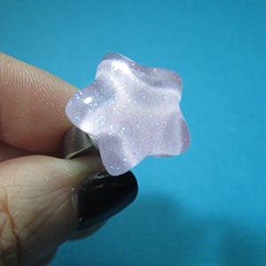 Puede incluir: Un anillo en forma de estrella con un dise&ntilde;o morado claro y brillante. La banda del anillo es plateada. El anillo se sostiene sobre un fondo azul.