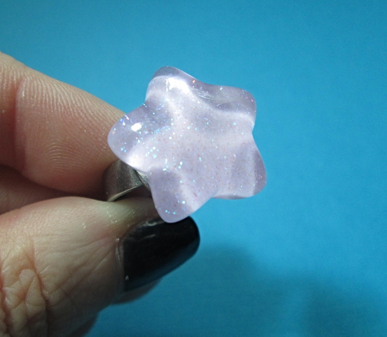 Puede incluir: Un anillo en forma de estrella con un dise&ntilde;o morado claro y brillante. La banda del anillo es plateada. El anillo se sostiene sobre un fondo azul.
