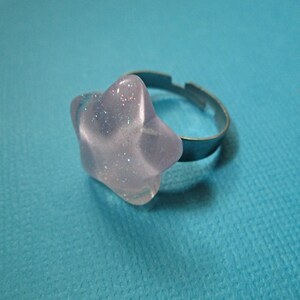 Puede incluir: Un anillo transparente en forma de estrella con detalles de purpurina plateada. El anillo tiene una banda ajustable plateada. La estrella mide aproximadamente 2,5 cm de di&aacute;metro. El anillo est&aacute; sobre un fondo azul.