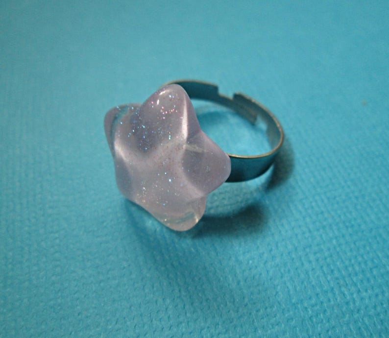 Puede incluir: Un anillo transparente en forma de estrella con detalles de purpurina plateada. El anillo tiene una banda ajustable plateada. La estrella mide aproximadamente 2,5 cm de di&aacute;metro. El anillo est&aacute; sobre un fondo azul.