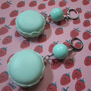 May include: Two mint green macaron-shaped keychains with silver-colored hardware. Each keychain has a round bead and a clasp. The background is pink with a pattern of red strawberries.