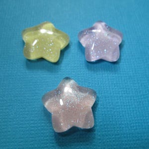 Puede incluir: Tres cuentas brillantes en forma de estrella en tonos amarillo, lavanda y transparente con purpurina iridiscente. Las cuentas est&aacute;n dispuestas sobre un fondo azul turquesa. Adecuadas para manualidades y bisuter&iacute;a.