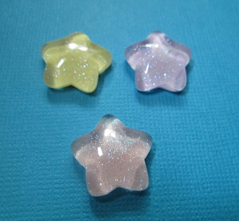 Puede incluir: Tres cuentas brillantes en forma de estrella en tonos amarillo, lavanda y transparente con purpurina iridiscente. Las cuentas est&aacute;n dispuestas sobre un fondo azul turquesa. Adecuadas para manualidades y bisuter&iacute;a.