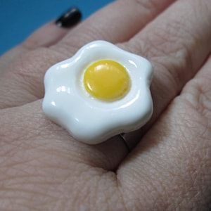 May include: A white and yellow fried egg ring. The ring features a glossy white egg white and a bright yellow yolk. The ring is a fun, novelty accessory.