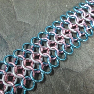 May include: A close-up of a chainmail bracelet with alternating blue, silver, and pink rings. The bracelet is made of metal and has a smooth, polished finish.