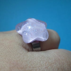 Puede incluir: Un anillo en forma de estrella de color lila claro con detalles de purpurina. El anillo tiene una banda plateada y est&aacute; sobre un fondo azul. La estrella mide aproximadamente 3,8 cm de ancho.
