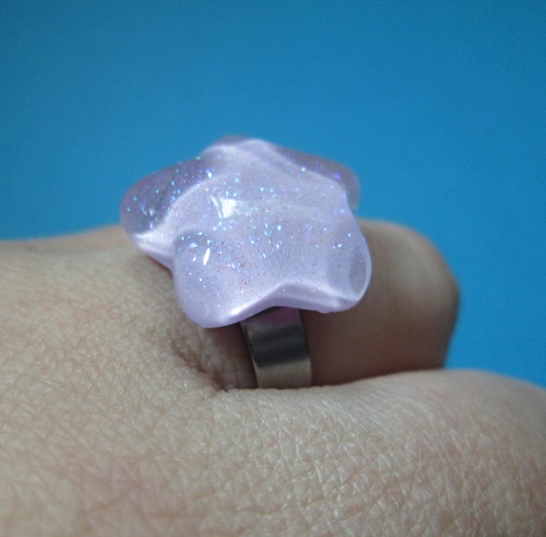 Puede incluir: Un anillo en forma de estrella de color lila claro con detalles de purpurina. El anillo tiene una banda plateada y est&aacute; sobre un fondo azul. La estrella mide aproximadamente 3,8 cm de ancho.