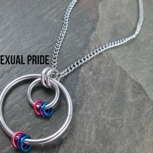 May include: A silver chain necklace with a pendant featuring two interlocking circles and a cluster of smaller rings in pink, blue, and purple. The text "Bisexual Pride" is visible at the top of the image.