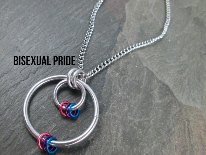 May include: A silver chain necklace with a pendant featuring two interlocking circles and a cluster of smaller rings in pink, blue, and purple. The text "Bisexual Pride" is visible at the top of the image.