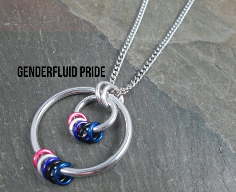 May include: A silver chain necklace with a pendant featuring two interlocking circles. The smaller circle is filled with small, colorful rings in the colors of the genderfluid pride flag: pink, white, blue, black, and blue. The text "GENDERFLUID PRIDE" is visible at the top of the image.