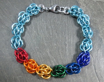 May include: A rainbow colored chainmail bracelet with a silver clasp. The bracelet is made of small metal rings that are linked together in a pattern. The colors of the bracelet are blue, green, yellow, orange, and red.