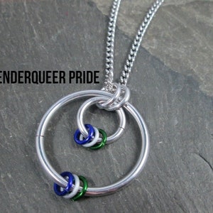 May include: A silver chain necklace with a large silver ring pendant. The ring has three smaller rings inside it, each with a different color: blue, white, and green. The text "GENDERQUEER PRIDE" is visible at the top of the image.