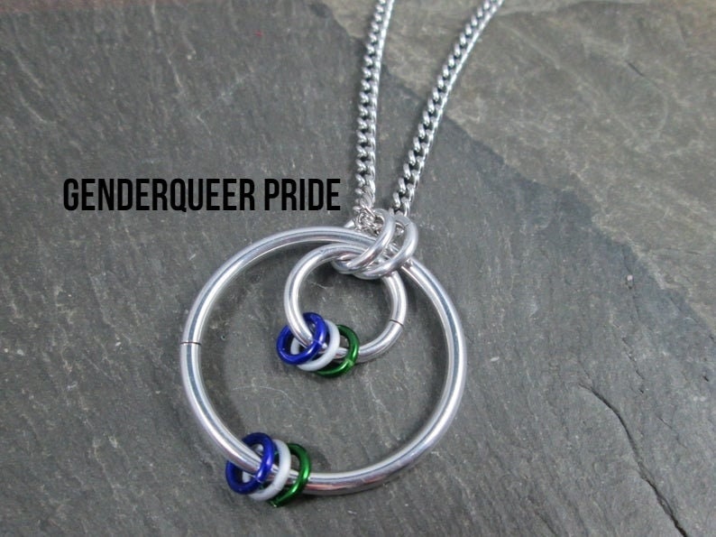 May include: A silver chain necklace with a large silver ring pendant. The ring has three smaller rings inside it, each with a different color: blue, white, and green. The text "GENDERQUEER PRIDE" is visible at the top of the image.
