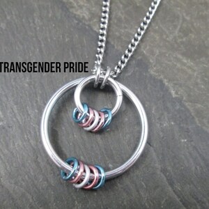 May include: A silver chain necklace with a silver circle pendant. The pendant has a smaller circle inside with a series of smaller rings in blue, pink, and white colors. The text "TRANSGENDER PRIDE" is visible at the top of the image.