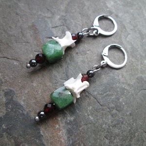 May include: A pair of earrings with green gemstone beads, white bone beads, and dark red and silver beads. The earrings have silver hoops.
