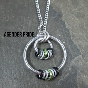May include: A silver necklace with a pendant featuring two interlocking circles. The circles are made of silver and are decorated with a cluster of small, colorful rings. The rings are black, green, and silver. The necklace is on a gray background. The text "AGENDER PRIDE" is visible in the image.