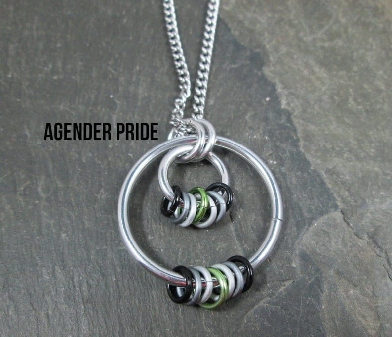 May include: A silver necklace with a pendant featuring two interlocking circles. The circles are made of silver and are decorated with a cluster of small, colorful rings. The rings are black, green, and silver. The necklace is on a gray background. The text "AGENDER PRIDE" is visible in the image.