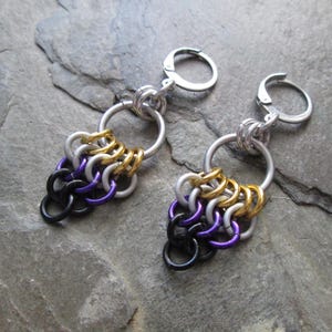 May include: A pair of dangle earrings with a chainmail design in black, purple, silver, and gold colors. The earrings are attached to silver hoop hooks.